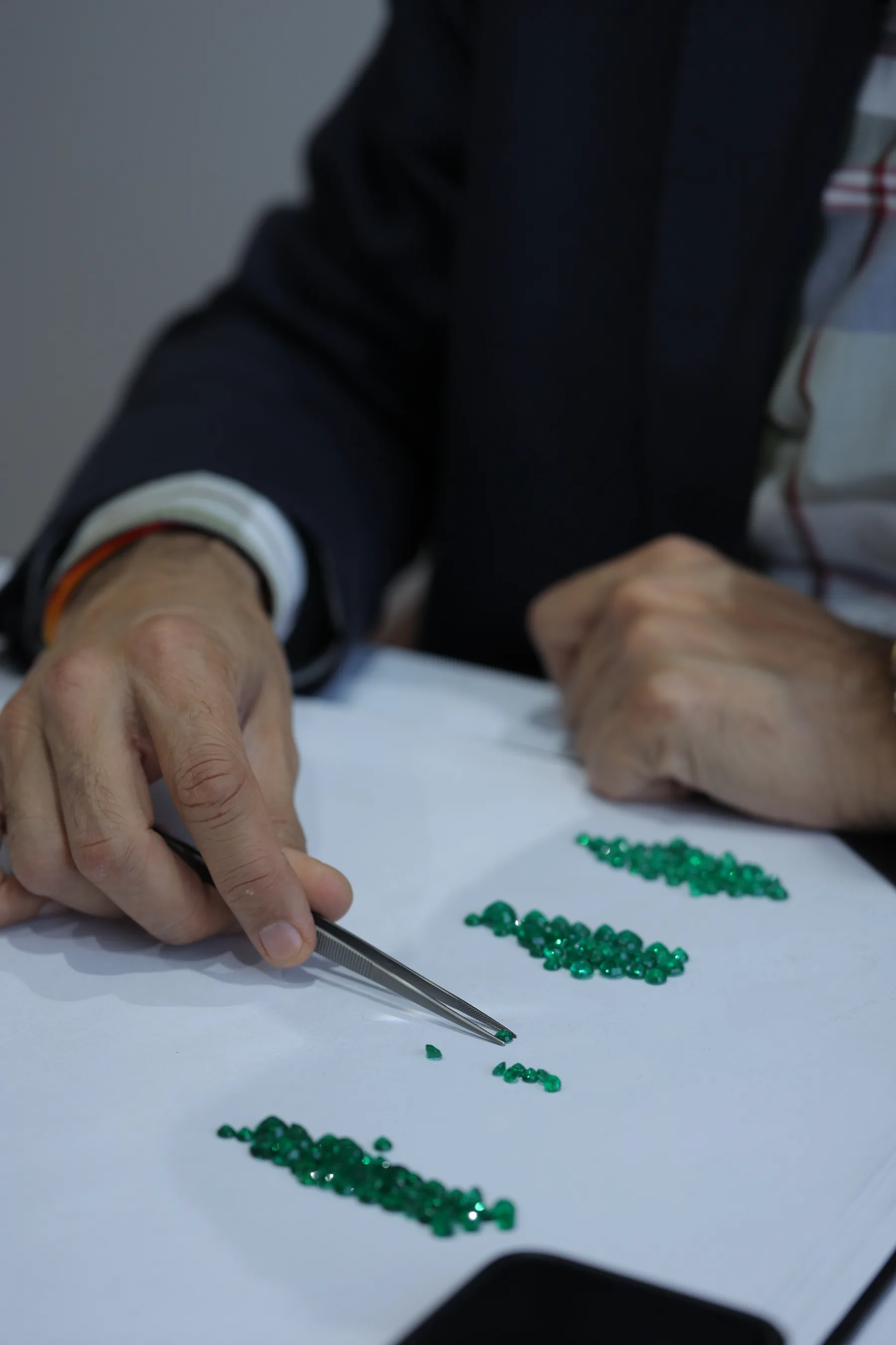 Overhead shot of a white tray filled with hundreds of small polished emeralds being sorted by color and clarity with tweezers